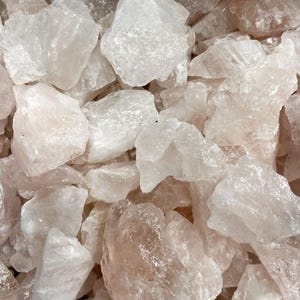 May include: A close-up shot of a pile of raw, rough-cut rose quartz crystals. The crystals are a pale pink color, with varying shapes and sizes. The light catches the facets of the stones, creating a sparkling effect. The overall theme is natural gemstones.