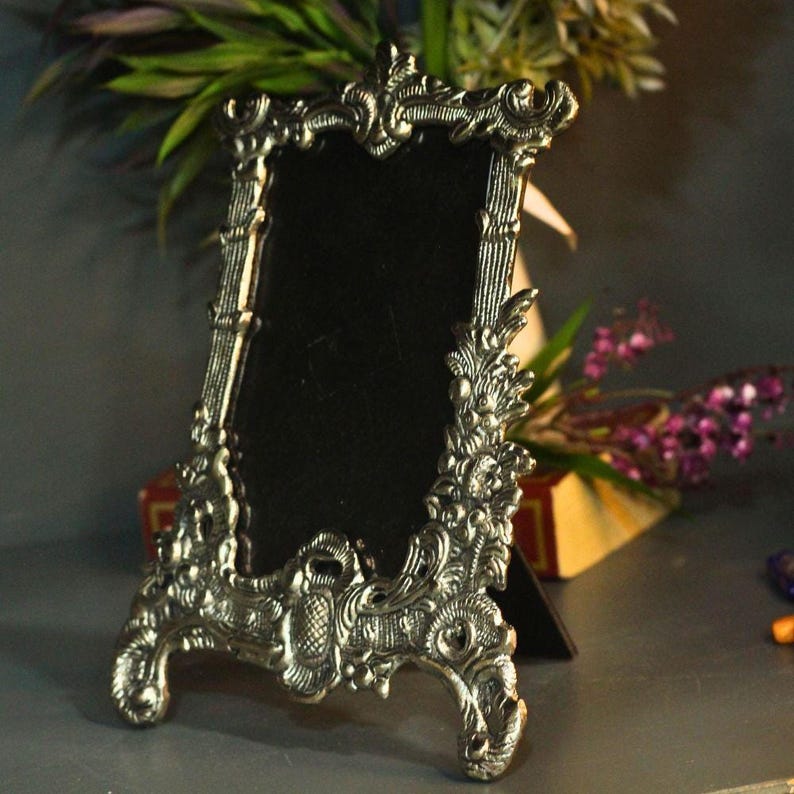 Vintage silver baroque-style picture frame with ornate floral scrollwork and an empty dark center on a tabletop.