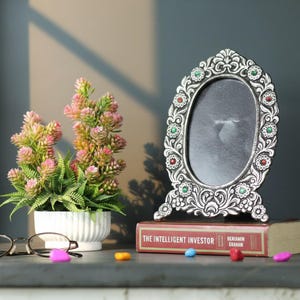 Puede incluir: Un marco de fotos ovalado plateado con detalles florales y de joyas. Se encuentra sobre un libro titulado "The Intelligent Investor". A la izquierda hay una pequeña planta en maceta con hojas rosas y verdes. Caramelos de colores esparcidos.