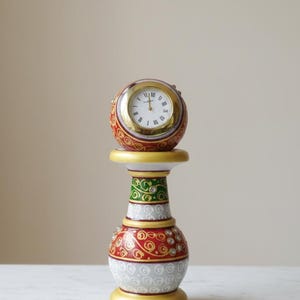 May include: A decorative table clock with a round face and Roman numerals. The clock sits atop a pedestal with red, green, and gold accents. The clock and pedestal are adorned with intricate patterns and embellishments.