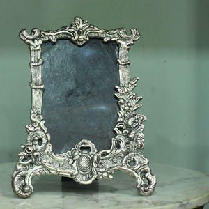 Vintage silver baroque-style picture frame with ornate floral scrollwork and an empty dark center on a tabletop.