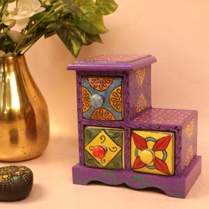 Może przedstawiać: Fioletowa, warstwowa drewniana skrzynia do przechowywania z czterema szufladami. Szuflady mają kolorowe, wzorzyste ceramiczne fronty z różnymi kolorami gałek. Skrzynia jest ozdobiona żółtymi kwiatowymi wzorami i kropkami.