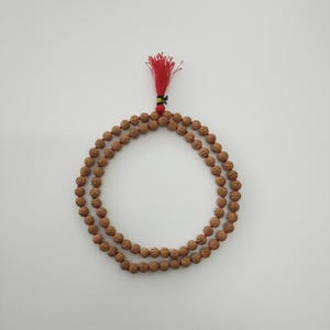 May include: A double-strand mala necklace with brown beads, arranged in a circular pattern. The necklace has a red tassel at the top, with a small green and yellow accent. The beads are a light brown colour.