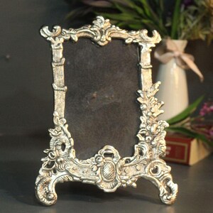 Vintage silver baroque-style picture frame with ornate floral scrollwork and an empty dark center on a tabletop.