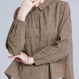 Può includere: Camicia marrone con pois bianchi. La camicia a maniche lunghe ha un colletto classico e un orlo leggermente asimmetrico. La modella indossa pantaloni bianchi e un braccialetto dorato. La camicia è abbottonata.