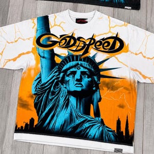 Puede incluir: Una camiseta blanca de manga corta con un gran estampado gráfico de la Estatua de la Libertad en tonos azules sobre un cielo naranja con rayos amarillos. La palabra GODSPEED está impresa en letras negras estilizadas sobre la estatua.