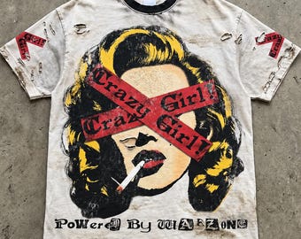 May include: A distressed off-white t-shirt with a black crew neck, featuring a graphic of a person with yellow hair smoking a cigarette. A red "Crazy Girl!" banner crosses the eyes. The text "Powered By WARZONE" appears at the bottom.