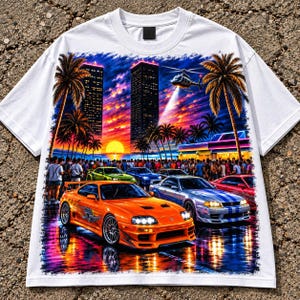 May include: A white short-sleeve t-shirt featuring a vibrant graphic print of a sunset cityscape with palm trees, a helicopter, and a crowd of people. In the foreground, a bright orange sports car and a silver sports car are parked on a wet surface.