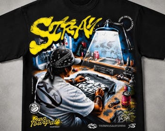 May include: A black t-shirt featuring a large yellow graphic that reads STRAY. The design depicts a person wearing a bandana and a white shirt, working at a desk under a lamp, with the text Own Your Path and other small details printed.