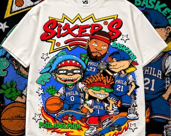 May include: A cream-colored short-sleeve t-shirt featuring a graphic print of cartoon characters wearing blue Philadelphia basketball jerseys. The design includes the text "SIXER'S" in red, "PHILADELPHIA" in green, and "BASKETBALL" in green, with basketballs and stars.