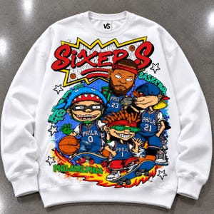 Puede incluir: Una sudadera blanca de cuello redondo con un estampado gr&aacute;fico colorido de cuatro personajes de dibujos animados con camisetas de baloncesto azules de Filadelfia numeradas 0, 23, 21 y 1. El texto SIXERS est&aacute; impreso en rojo arriba.