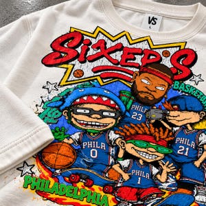 Puede incluir: Una sudadera de cuello redondo color crema con un estampado gr&aacute;fico de cuatro personajes de dibujos animados con camisetas de baloncesto azules de Filadelfia con los n&uacute;meros 0, 1, 21 y 23. El texto dice SIXER'S y PHILADELPHIA.