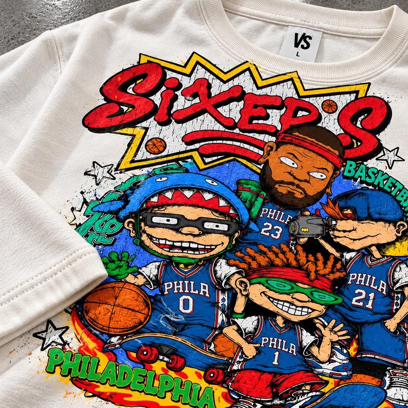 Puede incluir: Una sudadera de cuello redondo color crema con un estampado gr&aacute;fico de cuatro personajes de dibujos animados con camisetas de baloncesto azules de Filadelfia con los n&uacute;meros 0, 1, 21 y 23. El texto dice SIXER'S y PHILADELPHIA.