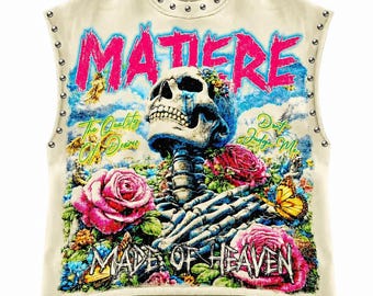 Puede incluir: Una camiseta sin mangas de color crema con tachuelas plateadas a lo largo del cuello y las sisas. El gráfico presenta una calavera, rosas rosas, una mariposa y el texto MATIERE, The Quality Of Desires, Don't Judge Me, MADE OF HEAVEN.
