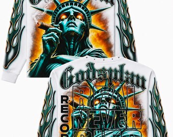May include: A white long-sleeve sweatshirt featuring a graphic of the Statue of Liberty smoking a cigar. The text reads Godsplans, Real Never Dies, and Records World.Tour. The sleeves are decorated with green and black flame patterns.