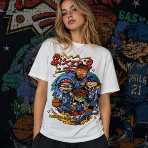 Puede incluir: Una camiseta blanca de manga corta con un estampado gr&aacute;fico colorido de personajes de dibujos animados con camisetas de baloncesto azules de Filadelfia. El texto Sixers est&aacute; impreso en rojo sobre los personajes y Philadelphia en verde.