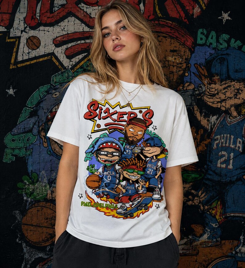 Puede incluir: Una camiseta blanca de manga corta con un estampado gr&aacute;fico colorido de personajes de dibujos animados con camisetas de baloncesto azules de Filadelfia. El texto Sixers est&aacute; impreso en rojo sobre los personajes y Philadelphia en verde.