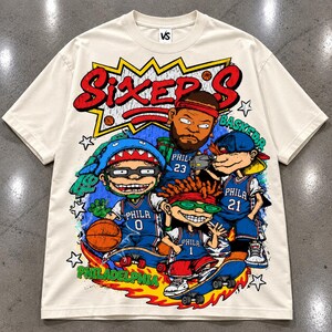 Puede incluir: Una camiseta de manga corta color hueso con un gran estampado gr&aacute;fico que incluye el texto "SIXER'S" y "PHILADELPHIA". El dise&ntilde;o muestra cuatro personajes de dibujos animados con camisetas de baloncesto azules numeradas 0, 1, 21 y 23.