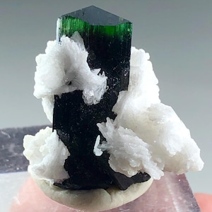 May include: A dark green tourmaline crystal with a hexagonal top, accented by white, fluffy mineral formations. The tourmaline is set on a light-colored base, showcasing its natural beauty. A striking mineral specimen.
