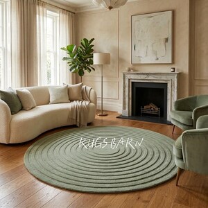 May include: A sage green circular rug with a concentric pattern is the focal point of a living room. The rug sits on a wooden floor, complemented by a curved sofa, armchairs, and a fireplace. The text "RUGS BARN" is displayed on the rug.