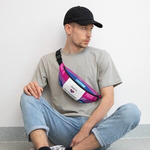 May include: A fanny pack with a bi pride flag design. The bag features pink, purple, blue, and white panels with the text "Pride Is For Everyone." The bag is worn across the chest. The person in the image is wearing a black cap, a gray t-shirt, and blue jeans.