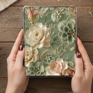 May include: A square, clear case featuring a floral design with raised details. The design includes cream, pink, and green flowers on a pale green background. The case is held by hands with dark red nail polish.