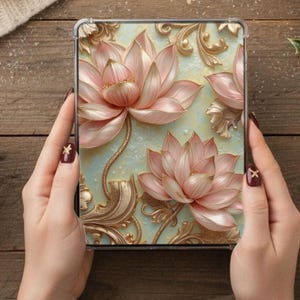 May include: A clear phone case featuring a 3D floral design with pink lotus flowers and gold accents. The background is a light blue color. The case is held by hands with dark red nail polish.