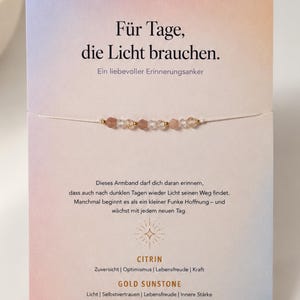 Könnte beinhalten: Ein zartes Armband mit kleinen, facettierten, mehrfarbigen Edelsteinperlen an einer dünnen weißen Schnur, befestigt auf einer rechteckigen Karte mit weichem Farbverlauf. Die Karte enthält deutschen Text, darunter CITRIN, GOLD SUNSTONE und BERGKRISTALL, ein Sternsymbol sowie den Schriftzug EUCH IM FOKUS am unteren Rand.