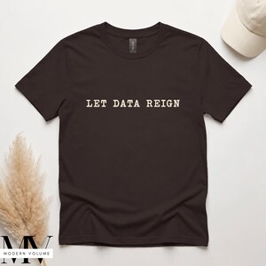 Pode incluir: Uma t-shirt de manga curta castanho-escura com o texto LET DATA REIGN impresso numa fonte serifada de cor creme claro no centro do peito. A peça tem um decote redondo clássico e um corte padrão, apresentada sobre uma superfície lisa.
