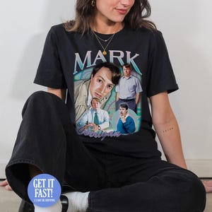 Peut inclure: T-shirt noir avec un graphisme rétro de Mark Corrigan de la sitcom britannique Peep Show. Le design comprend plusieurs images du personnage et le nom "MARK" dans une police vintage. Le t-shirt est porté.