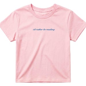 May include: A light pink t-shirt with the phrase "i'd rather be reading" in a cursive, navy blue font. The shirt has short sleeves and a crew neck. The fabric appears to be soft and comfortable.