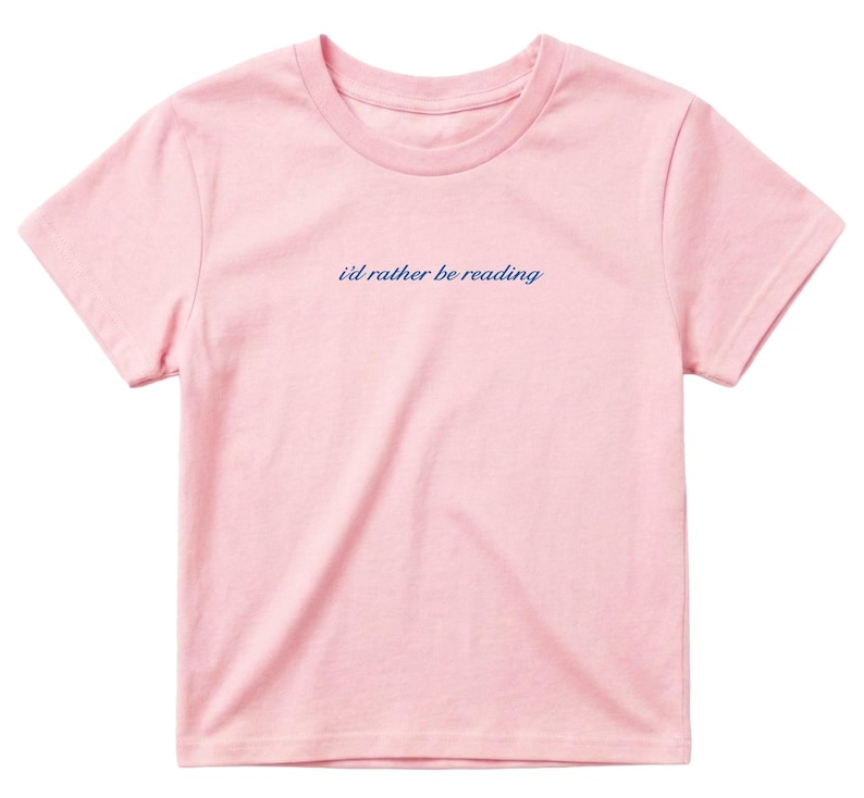May include: A light pink t-shirt with the phrase "i'd rather be reading" in a cursive, navy blue font. The shirt has short sleeves and a crew neck. The fabric appears to be soft and comfortable.