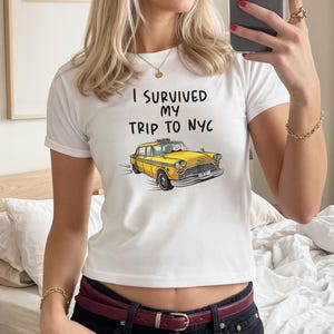 May include: White t-shirt featuring the text "I Survived My Trip To NYC" and a cartoon yellow taxi cab graphic. The shirt has short sleeves and a crew neck. The person is wearing a gold bracelet and rings.