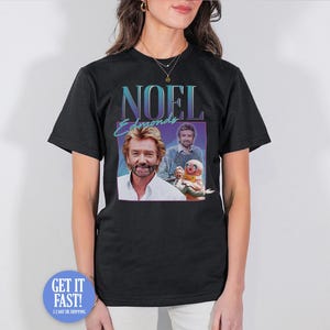 May include: Black t-shirt featuring a retro design with the name "NOEL EDMONDS" in teal and purple. The shirt includes two images of a man, one in a white shirt and the other in a sweater, with a puppet. The shirt is worn by a person.