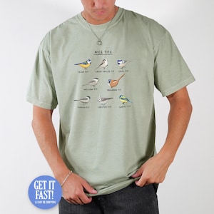May include: A sage green t-shirt featuring the phrase "NICE TITS" above illustrations of different bird species. Each bird is labelled with its common name, including "Blue Tit" and "Long Tailed Tit".
