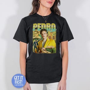 Peut inclure: T-shirt noir avec un graphisme de Pedro Pascal en blazer jaune, avec le texte "PEDRO Pascal" en jaune et bleu. Le design comprend plusieurs images de l'acteur. Le t-shirt est porté par une personne.