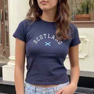 Peut inclure: T-shirt bleu marine avec le mot "SCOTLAND" en blanc, arqué sur la poitrine, et un petit drapeau écossais en dessous. Le t-shirt a des manches courtes et un col rond.