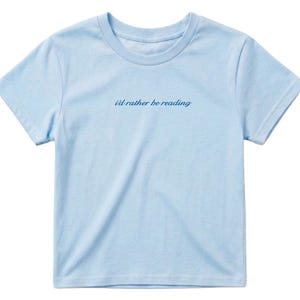 May include: A light blue t-shirt with the phrase "i'd rather be reading" in a cursive font. The shirt has short sleeves and a crew neck. The fabric appears to be soft and lightweight, suitable for casual wear.