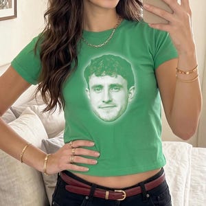 Può includere: Un crop top verde con un ritratto di un uomo. La maglietta ha maniche corte e girocollo. La persona nell'immagine indossa gioielli d'oro e pantaloni neri con cintura rossa.