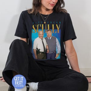 Può includere: T-shirt nera con una grafica di due uomini e la scritta "Hitchcock & Scully" in oro e blu. La t-shirt è indossata da una persona seduta per terra.