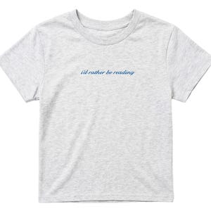 May include: A light gray t-shirt with the text "i'd rather be reading" in a cursive, navy blue font. The shirt has short sleeves and a crew neck. The fabric appears to be a soft cotton blend.