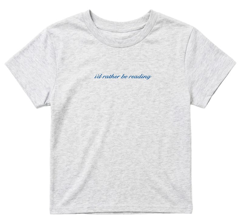 May include: A light gray t-shirt with the text "i'd rather be reading" in a cursive, navy blue font. The shirt has short sleeves and a crew neck. The fabric appears to be a soft cotton blend.