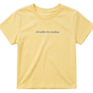 May include: A yellow t-shirt with the phrase "I'd rather be reading" in a dark blue script across the chest. The shirt has a crew neck and short sleeves, and is made of a soft, lightweight material.