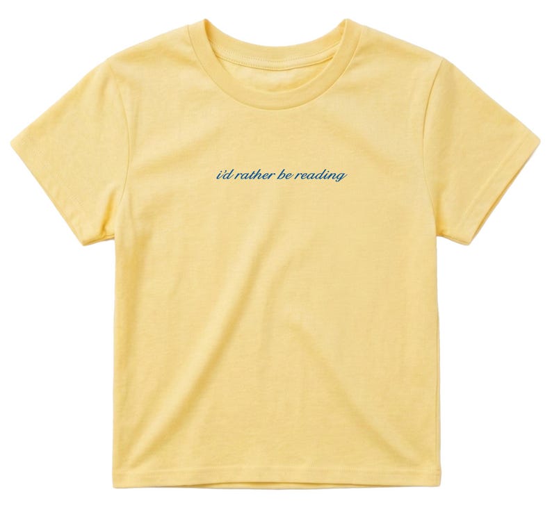 May include: A yellow t-shirt with the phrase "I'd rather be reading" in a dark blue script across the chest. The shirt has a crew neck and short sleeves, and is made of a soft, lightweight material.
