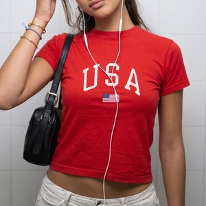 May include: A red crop top with the word "USA" in white block letters and a small American flag graphic. The top is paired with white trousers and a black shoulder bag. White earbuds are visible.