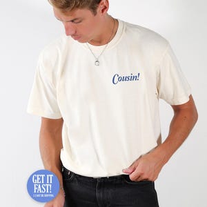 Op de afbeelding: Crèmekleurig T-shirt met het woord "Cousin!" in blauw gedrukt. Het shirt is een model met korte mouwen en een ronde hals. Het model draagt een zwarte jeans en een zilveren ketting.