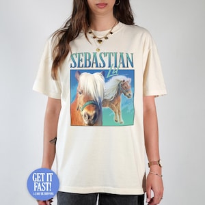May include: A cream-coloured t-shirt with a graphic of two horses and the text "SEBASTIAN 74" in blue and green. The shirt has short sleeves and a relaxed fit.