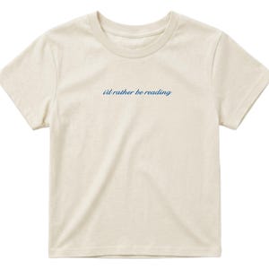 May include: Cream-colored t-shirt with short sleeves and a crew neck. The phrase "i'd rather be reading" is printed in a cursive, navy blue font across the chest. The shirt is made of a soft, lightweight fabric.
