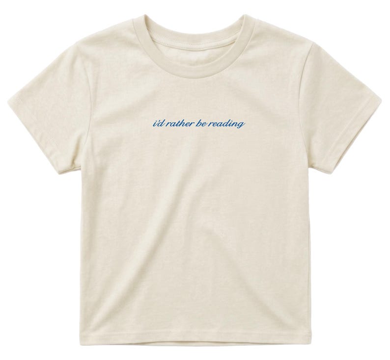 May include: Cream-colored t-shirt with short sleeves and a crew neck. The phrase "i'd rather be reading" is printed in a cursive, navy blue font across the chest. The shirt is made of a soft, lightweight fabric.