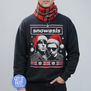 以下が含まれることがあります： 「snowasis」の文字と、サンタ帽をかぶった2人の人物のピクセル化されたグラフィックが特徴の濃いネイビーブルーのスウェットシャツ。デザインにはイギリスの国旗と雪の結晶が含まれています。人物は赤と緑のチェック柄のスカーフを着用しています。